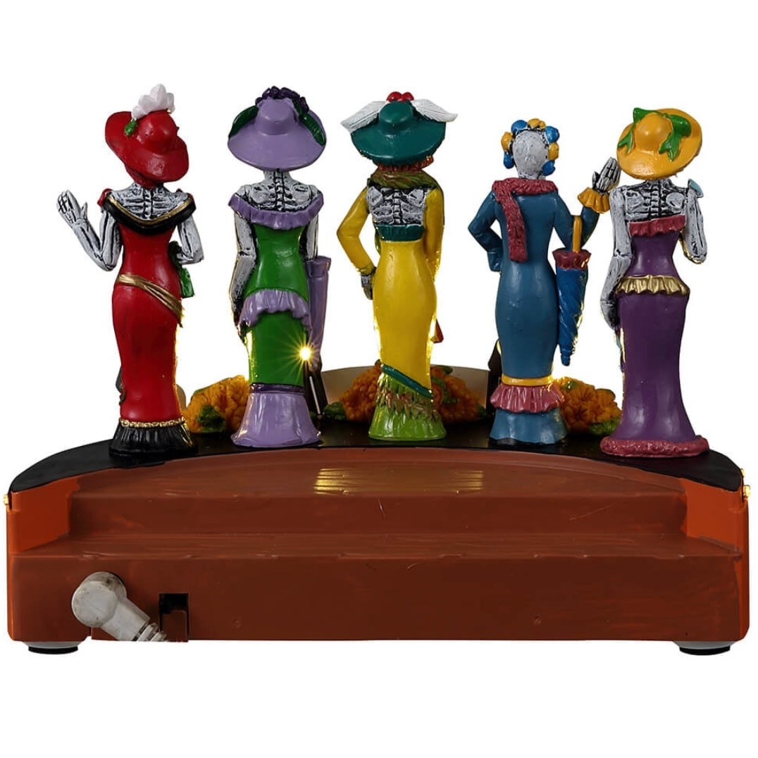 Resin accent for a Lemax Spooky Town display. Designed to look like five skeletons dressed in gaudy, brightly colored costumes. There are signs that read "Nuevo," "Dia de los Muertos Fashion Show" and "Elegante!" Designed to look like they're on stage at a fashion show. There are sugar skull accents and flowers on the stage.