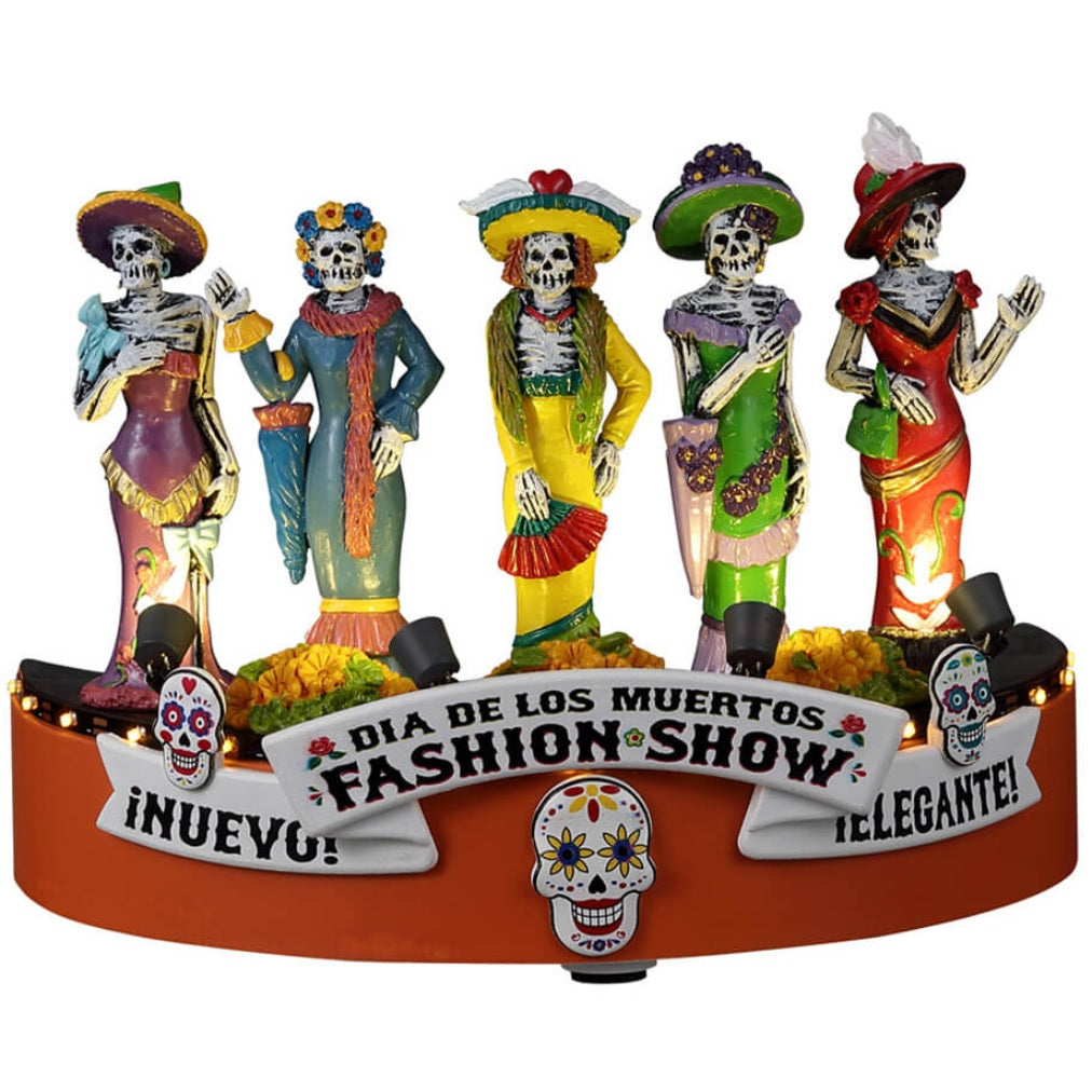 Resin accent for a Lemax Spooky Town display. Designed to look like five skeletons dressed in gaudy, brightly colored costumes. There are signs that read "Nuevo," "Dia de los Muertos Fashion Show" and "Elegante!" Designed to look like they're on stage at a fashion show. There are sugar skull accents and flowers on the stage.