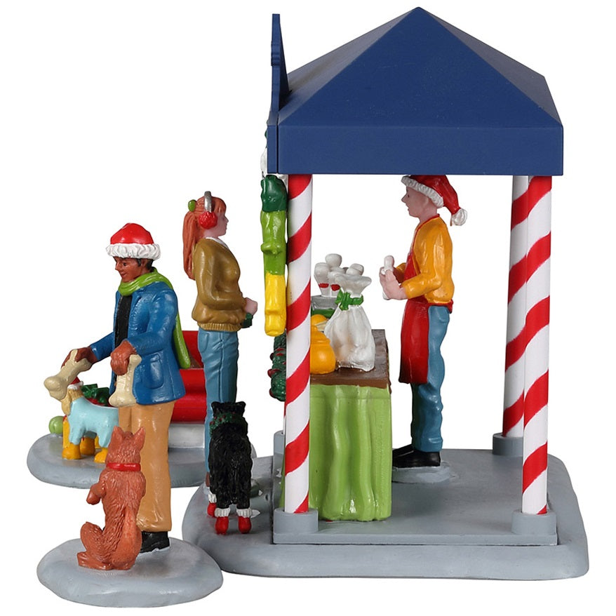 Resin table accent for a Lemax display. Designed to look like a booth that sells pet treats. It's decorated for Christmas. There's a woman handing her dog a bone while another is purchasing some food for her dog who is wearing red booties. There are two dogs wearing Santa hats in a red sleigh posing for pictures.