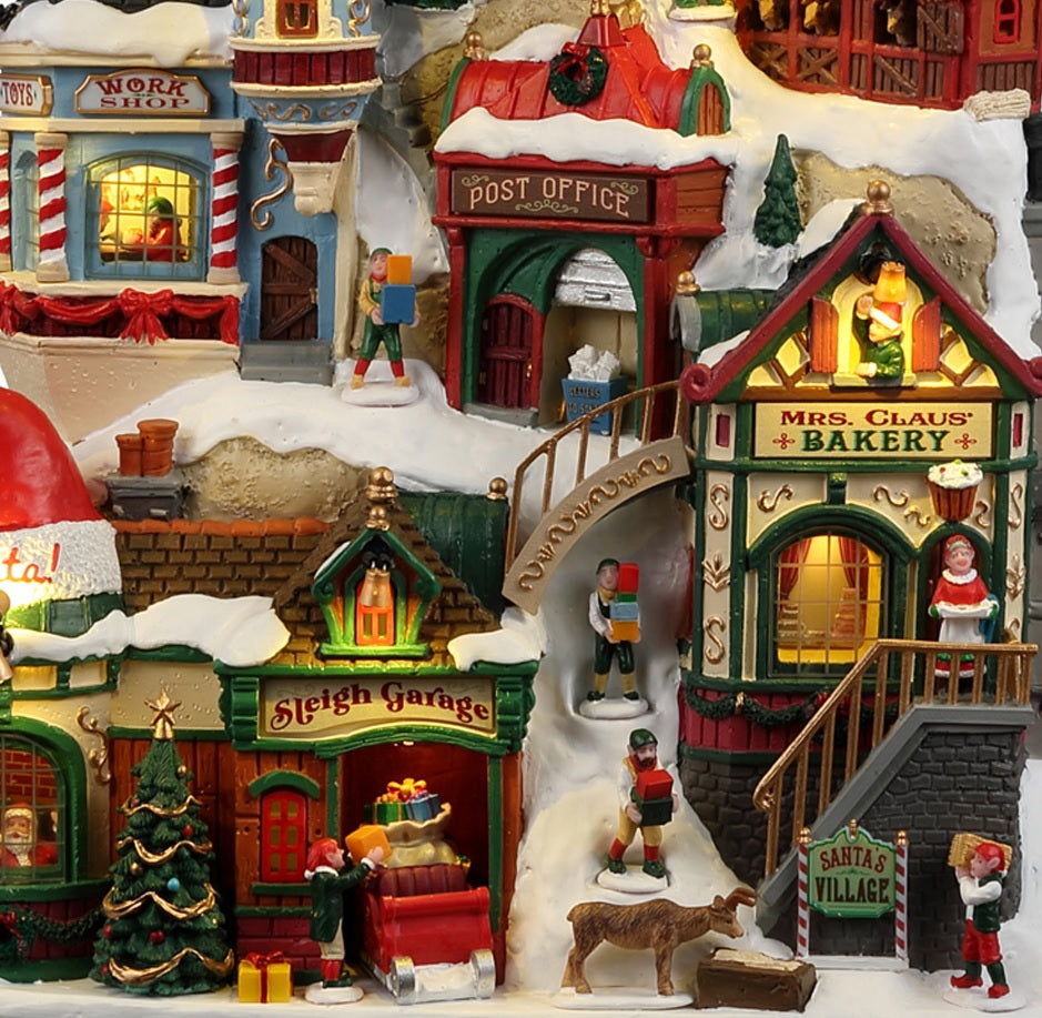 Resin building facade for a Lemax display. Designed to look like a bunch of store fronts decorated for Christmas. The roofs and the sidewalks are covered in snow. There are elves and reindeer and people shopping. There's a window on the left side where you can see Santa inside. There's a work shop, a post office, a sleigh garage, a bakery and some stables.