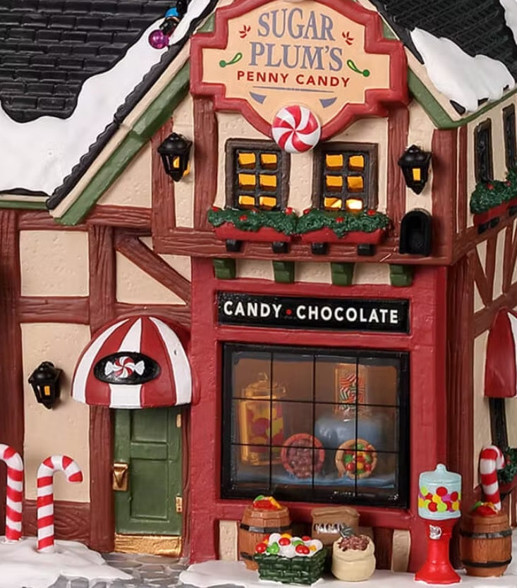 Porcelain lighted building for a Lemax display. Designed to look like a candy shop. There's snow on the roof and the sidewalk. Inside the window, you can see a bunch of candy on display. In front of the shop, there are peppermint candy canes, baskets of candy and a bubblegum machine.