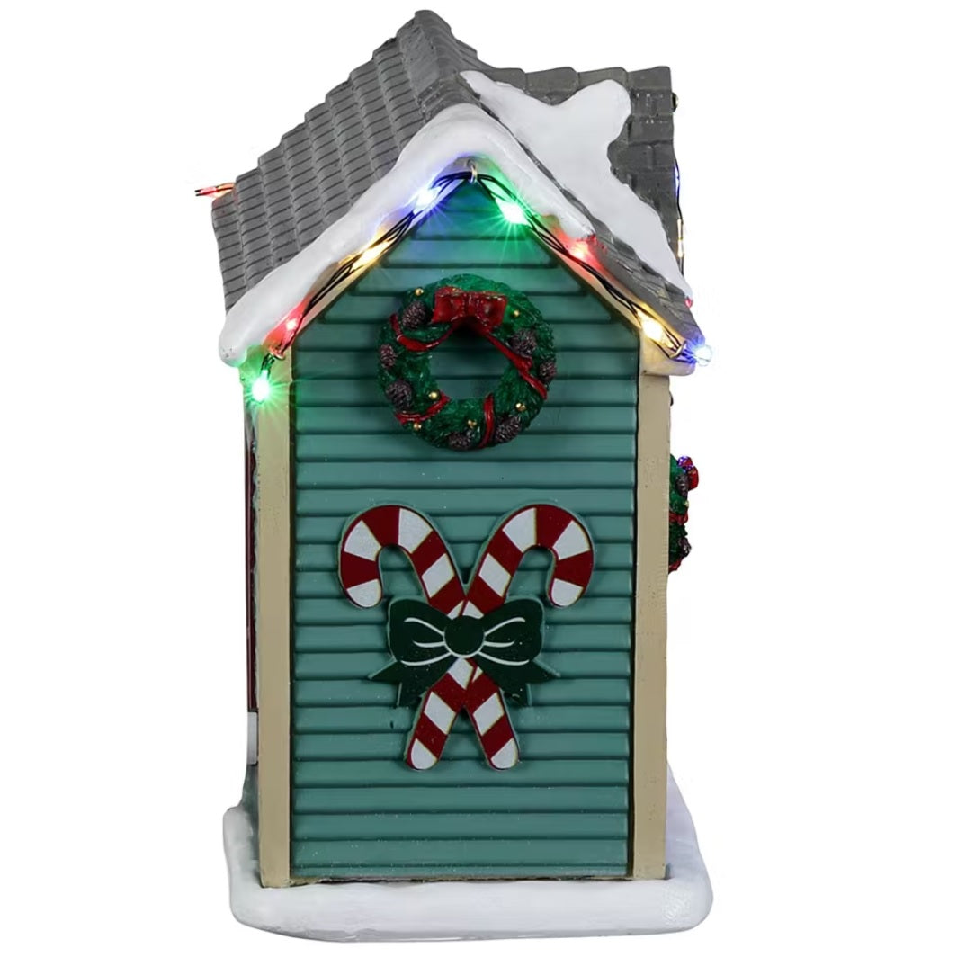 Resin village accent for a Lemax display. Designed to look like a turquoise shed with red barn doors and a gray roof. There's snow on the roof and multi-colored lights. There are wreaths on the doors and candy canes painted on the side. Inside the shed, you can see a woman decorating a wreath.