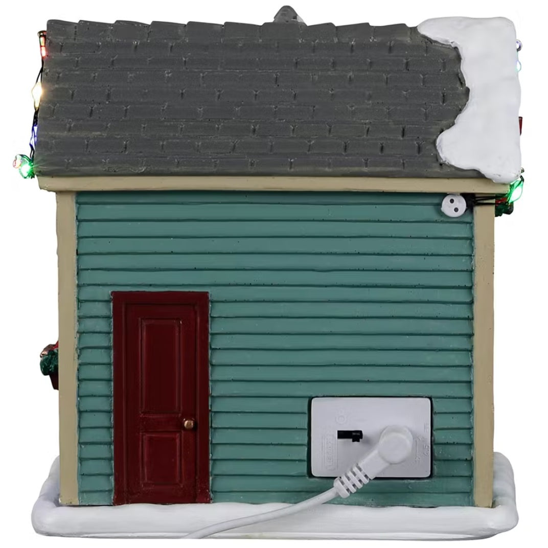 Resin village accent for a Lemax display. Designed to look like a turquoise shed with red barn doors and a gray roof. There's snow on the roof and multi-colored lights. There are wreaths on the doors and candy canes painted on the side. Inside the shed, you can see a woman decorating a wreath.