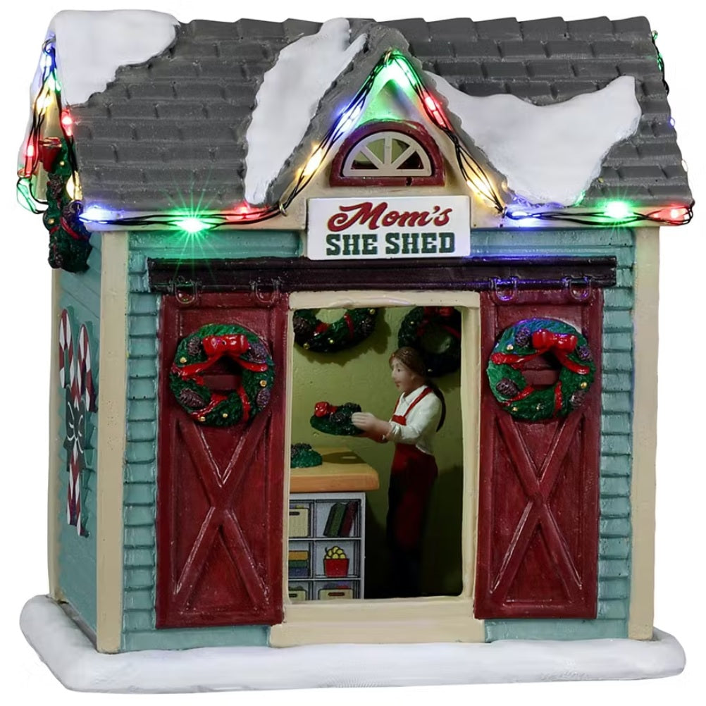 Resin village accent for a Lemax display. Designed to look like a turquoise shed with red barn doors and a gray roof. There's snow on the roof and multi-colored lights. There are wreaths on the doors and candy canes painted on the side. Inside the shed, you can see a woman decorating a wreath.