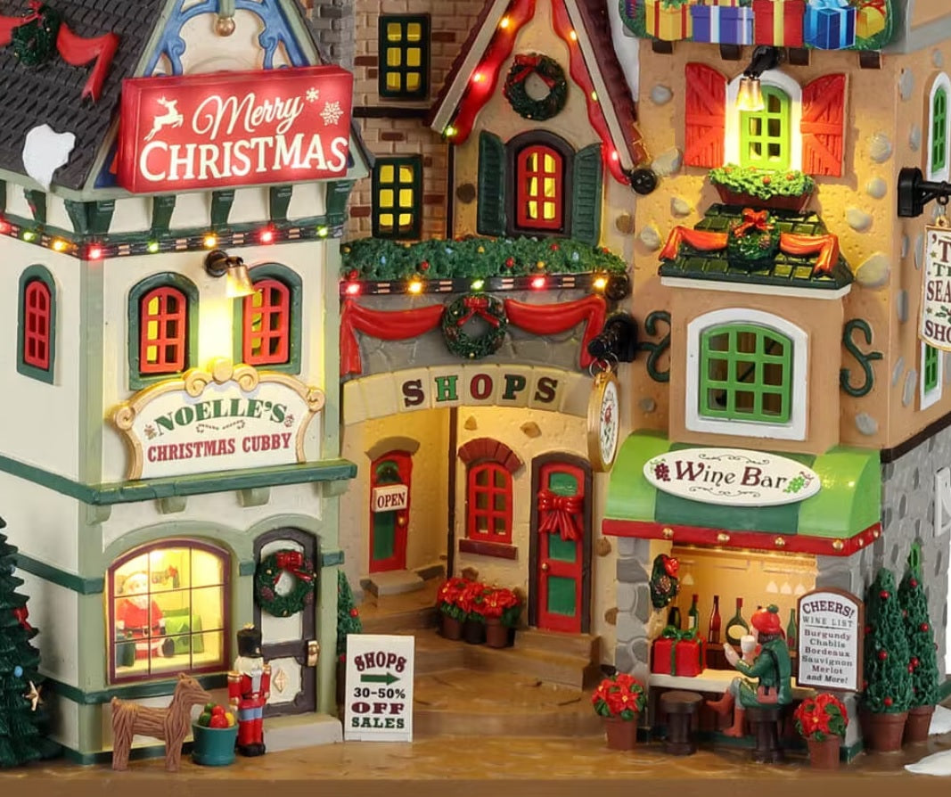 Polyresin building facade for a Lemax display. Designed to look like a collection of store fronts that are decorated for Christmas. Signs read "Merry Christmas," "Noelle's Christmas Cubby," "Shops 30-50% off sales," "Shops," "Wine Bar," "Cheers Wine List" and Tis the Season Shop."