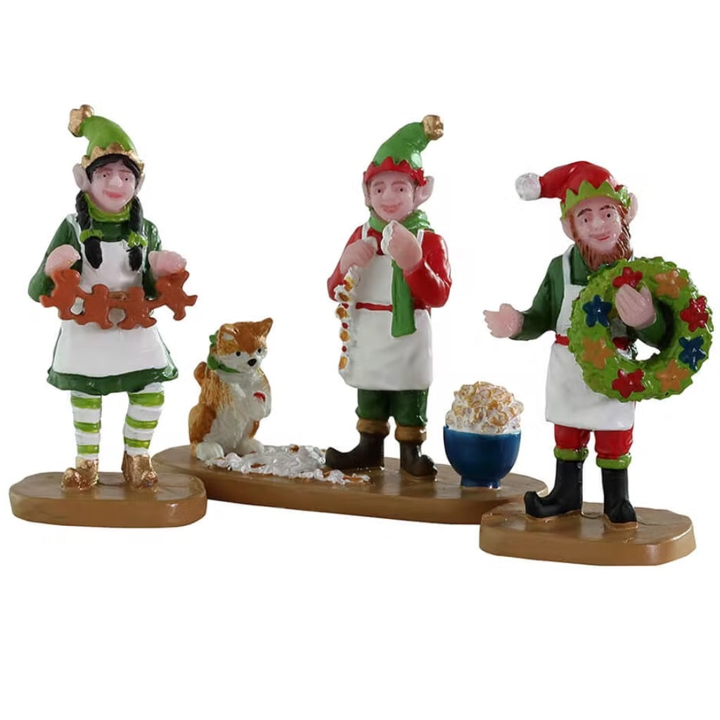 Resin figurines for a Lemax display. Designed to look like three elves creating decorations for Christmas. One is holding a garland made of gingerbread men, one elf is making a garland out of popcorn and another is holding a finished wreath. There's a dog sneaking some popcorn.