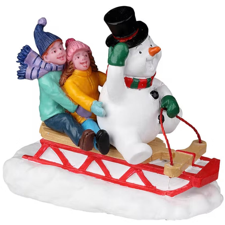 Resin figure for a Lemax Village tabletop display. Designed to look like two children and a snowman on a sled. They're all smiling. The snowman is wearing a top hat, green gloves and a red and green scarf. The kids are wearing winter clothes. The sled is red, white and brown.