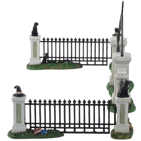 Polyresin village accessory for a Lemax display. Designed to look like an iron fence on patches of grass. There are four columns with witch hat and black cat statues on top. The gate reads "Witch's Gate" and there's a broom leaning against it.