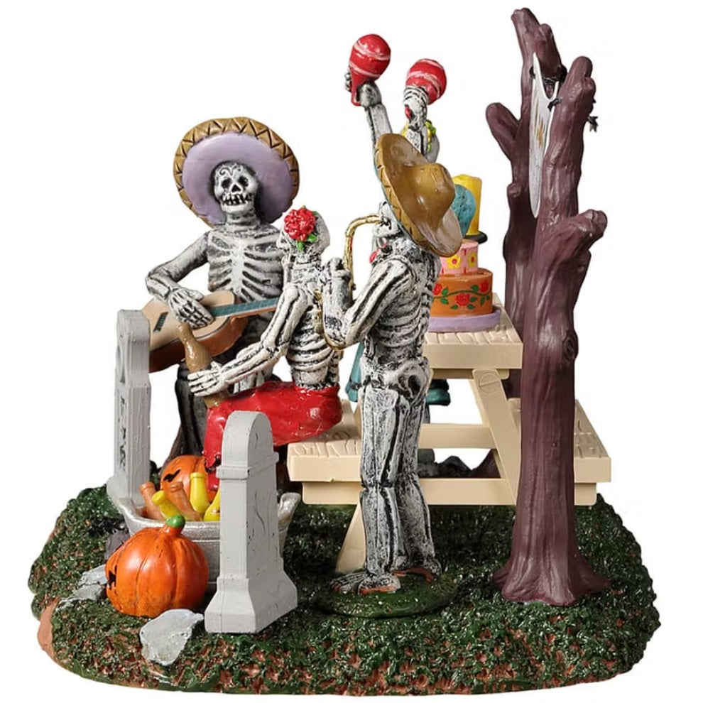 Resin village accent for a Spooky Town Lemax display. Designed to look like four skeletons enjoying a Day of the Dead celebration. There are tombstones, carved pumpkins and a banner hanging from the trees. There's a picnic table with a skull, a candle, a decorated cake and a rat on it.
