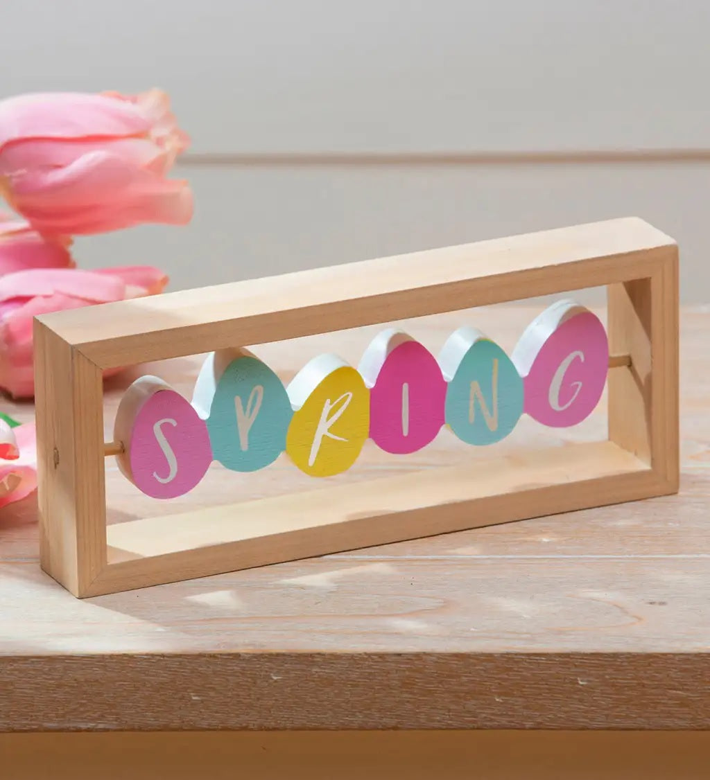 A rectangular wooden tabletop decoration made up of brightly colored eggs. The side that is shown here reads SPRING. The eggs are pink, light blue, yellow and bright pink