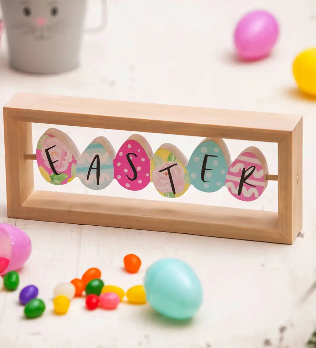 A rectangular wooden tabletop decoration made up of brightly colored eggs. The side that is shown here reads EASTER. Each egg has a different pattern on it. The colors are pink, light blue, green, pink, white, teal and yellow