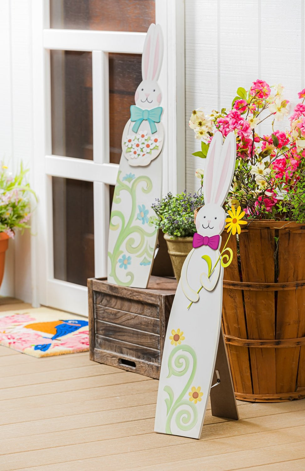 A porch decoration shaped like a tall bunny. One bunny is wearing a pink bow and holding a yellow flower and features two yellow flowers at the base. One bunny is wearing a teal bow and holding a bunch of white flowers and features four blue flowers at the base
