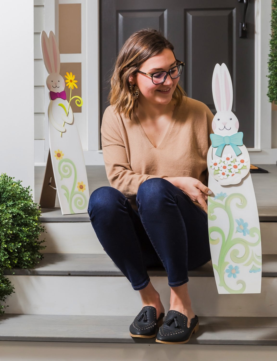 A porch decoration shaped like a tall bunny. One bunny is wearing a pink bow and holding a yellow flower and features two yellow flowers at the base. One bunny is wearing a teal bow and holding a bunch of white flowers and features four blue flowers at the base. Woman is sitting on the porch holding one of the decorations