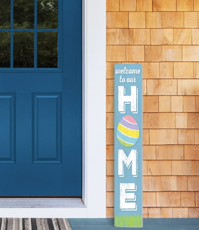 The vertical porch sign on a front porch next to a blue door. The sign reads "Welcome to our Home" and is perfect for Easter. The O in "home" is a multi-colored egg