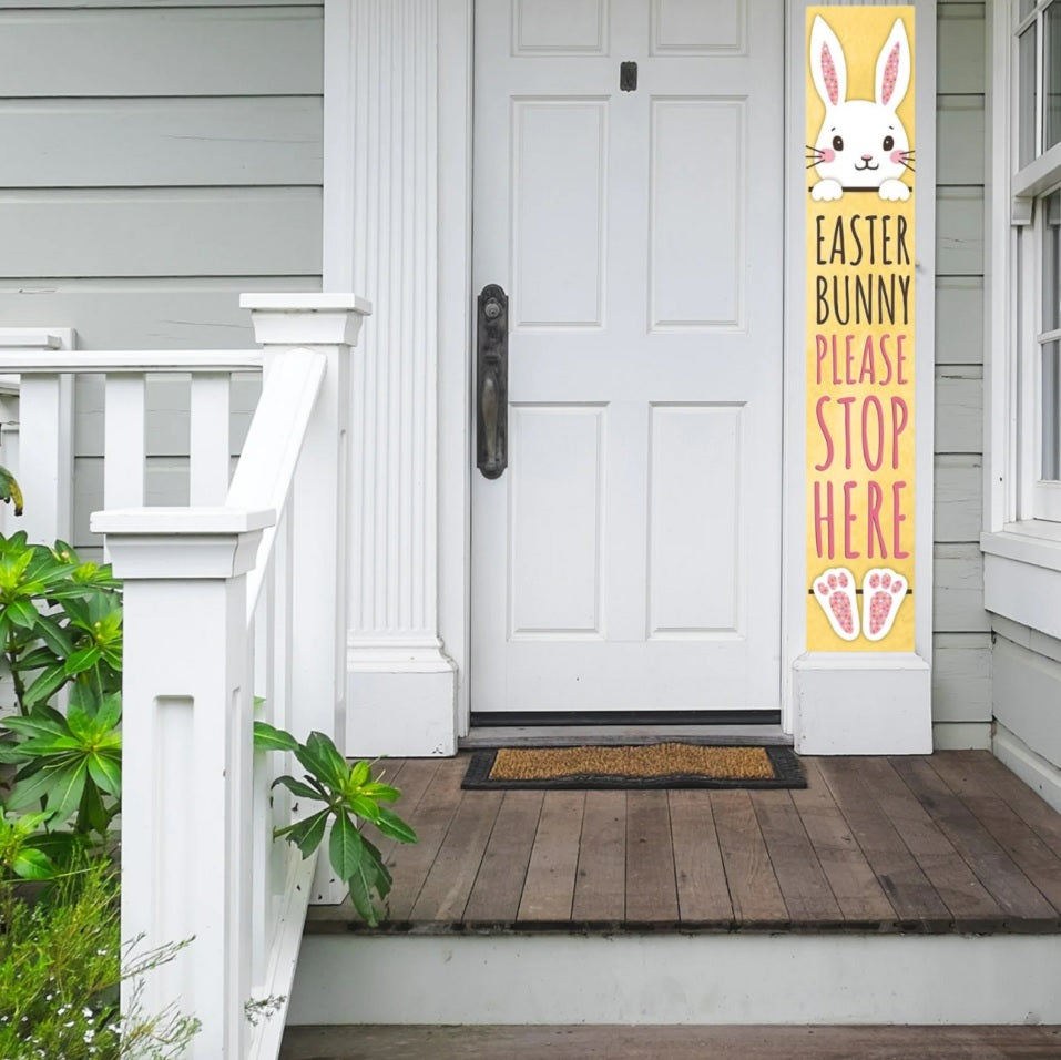 A vertical porch sign that reads "Easter Bunny Please Stop Here" on a yellow background. The top of a bunny is seen at the top and the bunny's feet are at the bottom