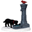 Polyresin figurine for a Lemax Village tabletop display. Designed to look like a black dog wearing a red collar drinking water out of a fountain. There's a red bird on top of the fountain. The ground is covered in snow