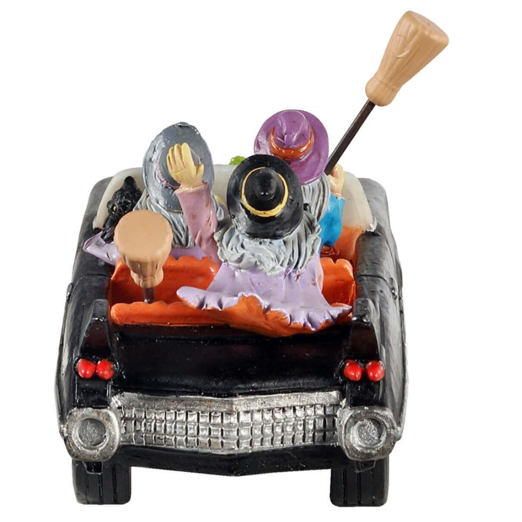 Polyresin figurine for a Lemax display. Designed to look like three witches out for a drive. They're all holding broomsticks, and two of them are holding black cats. There's a green toad on the front of the car.