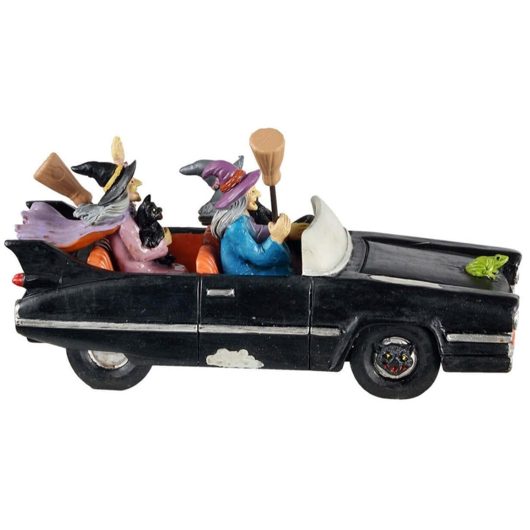 Polyresin figurine for a Lemax display. Designed to look like three witches out for a drive. They're all holding broomsticks, and two of them are holding black cats. There's a green toad on the front of the car.
