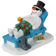 Polyresin figurine for a Lemax Village display. Designed to look like a snowman resting on a lounge chair. He is wearing a top hat, sunglasses and green and red shorts. He's holding a drink in one hand. His boots are off to the side of the chair. There's a book on the ground that reads "The Life of Frosty"
