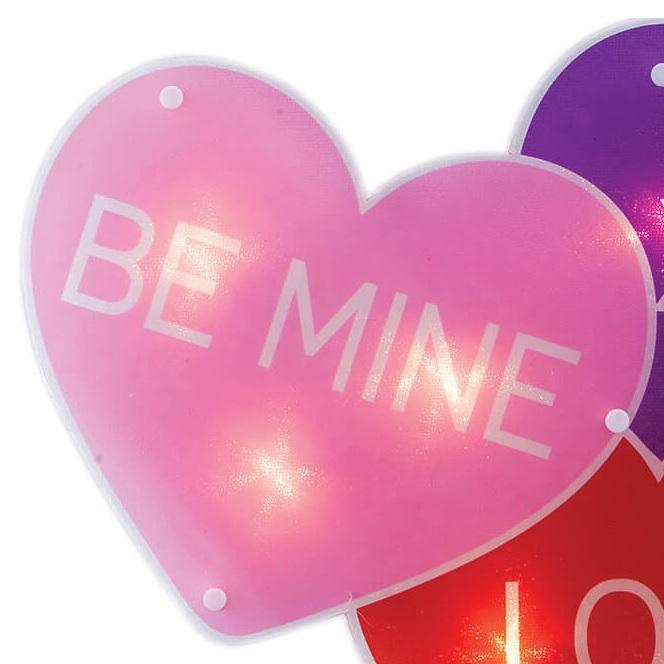 A light up decoration for Valentine's Day designed to look like three conversation hearts. One is purple and reads "XOXO." One is pink and reads "BE MINE." The other is red and reads "LOVE."
