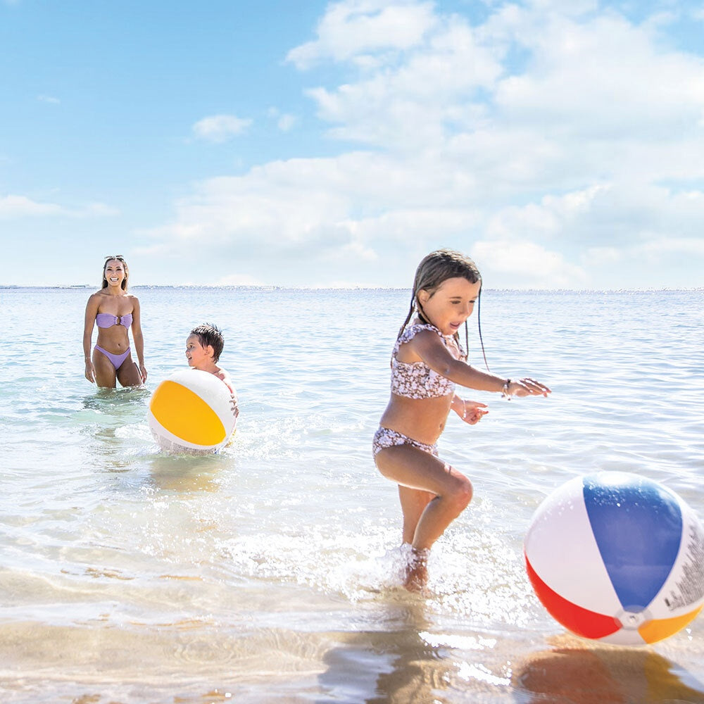 This is a lifestyle image showing two young kids and a mom frolicking in the ocean. They both have multi-colored beach balls.