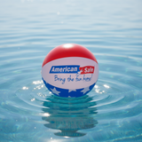 This is a lifestyle image showing a beach ball on top of the water. The beach ball is red, white and blue striped and reads "American Sale. Bring the fun home!"