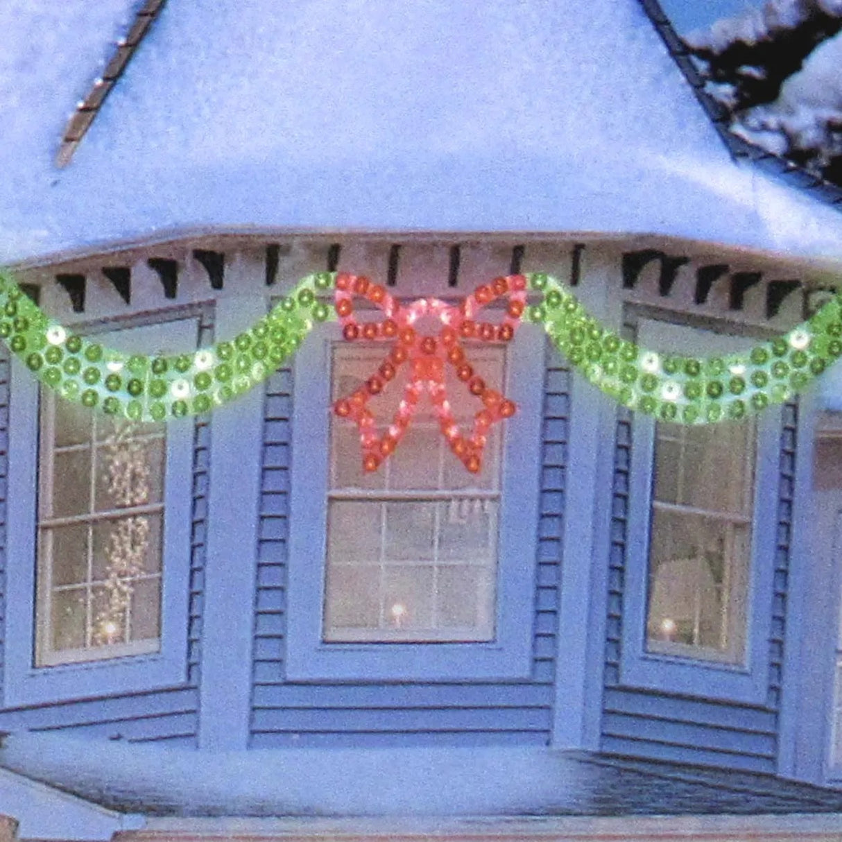 This is a lifestyle image showing the 6 foot long lighted Christmas decoration designed to look like green ribbon with a red bow in the center. In this image, it's across three windows at the front of a snowy house.