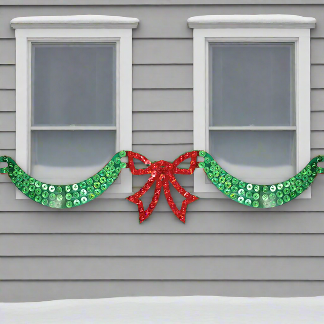 This is a lifestyle image showing the 6 foot long lighted Christmas decoration designed to look like green ribbon with a red bow in the center. In this image, it's across two windows at the front of a snowy house.