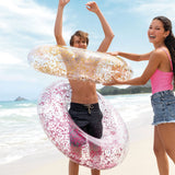 This is a lifestyle image showing a young girl putting two of these glitter inner tubes over a young boy on a beach.