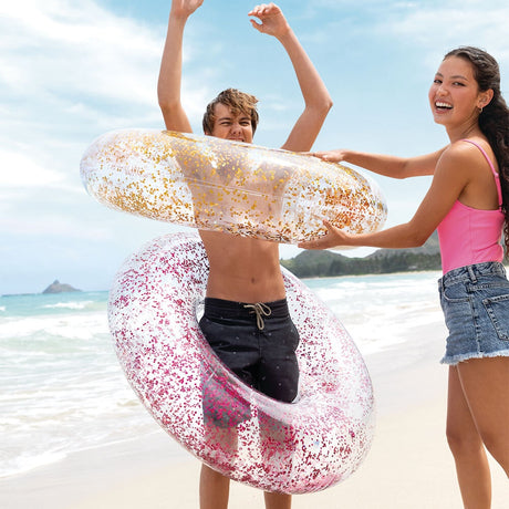 This is a lifestyle image showing a young girl putting two of these glitter inner tubes over a young boy on a beach.