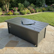 This is a lifestyle image showing what this fire pit table looks like on a stone patio in a grassy backyard.
