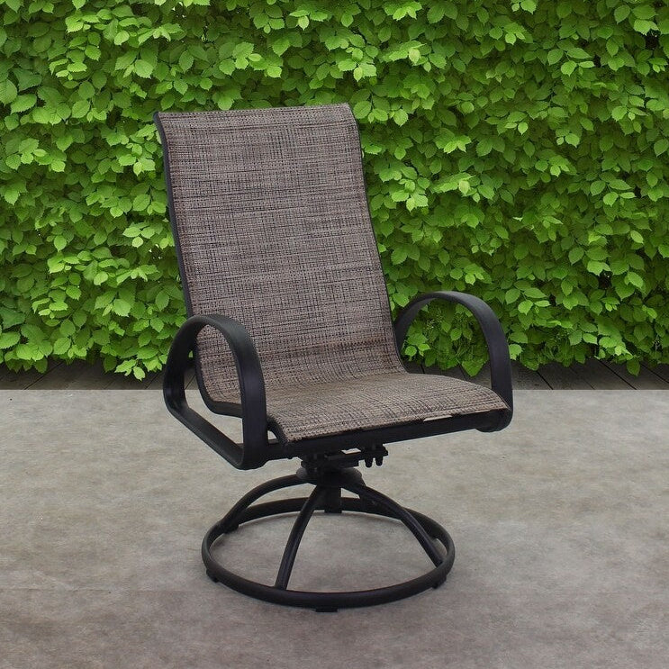 This is a lifestyle image showing this specific outdoor swivel rocker chair on a stone patio with a large, green bush behind it.