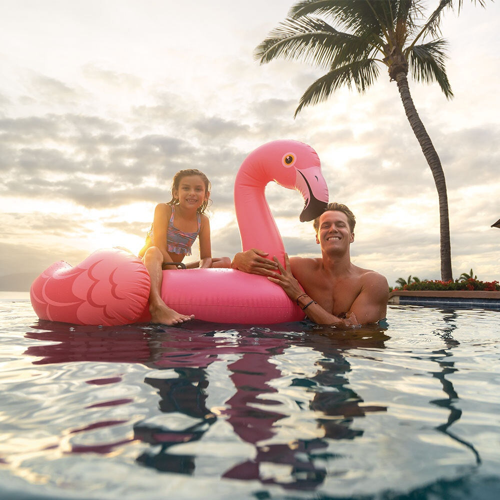 This is a lifestyle image showing a young girl sitting on this flamingo float in a pool while her dad holds the head of the float.