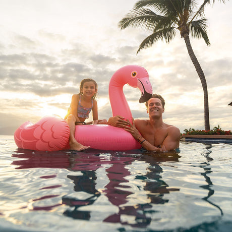 This is a lifestyle image showing a young girl sitting on this flamingo float in a pool while her dad holds the head of the float.
