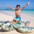 This is a lifestyle image showing a young boy kneeling on the inflatable turtle on a beach.