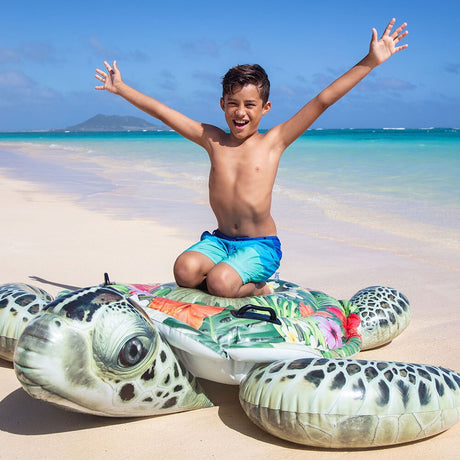This is a lifestyle image showing a young boy kneeling on the inflatable turtle on a beach.