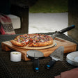 This is a lifestyle image showing off this Pizza BBQ Starter Kit in use. There's a pizza on the pizza stone and that is on the spatula. The pizza cutter and the grated spatula are resting next to it. There are plates on the table, as well.