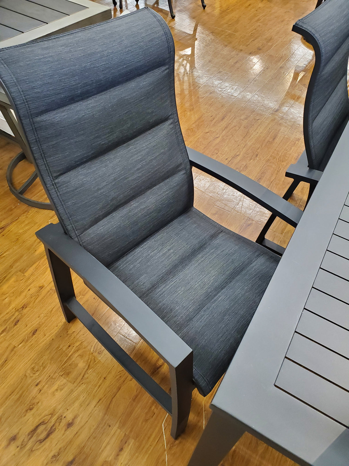 This is a lifestyle image showing the dining chair and part of the dining table. The frames of the chair and table are black. The padded sling fabric is a dark gray color.