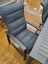 This is a lifestyle image showing the dining chair and part of the dining table. The frames of the chair and table are black. The padded sling fabric is a dark gray color.