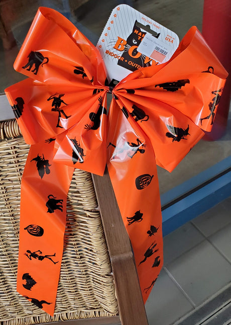 Orange bow with black figures on it. The figures include a skeleton, a pumpkin, a cat, a switch on a broom and a bat.
