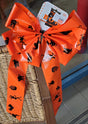 Orange bow with black figures on it. The figures include a skeleton, a pumpkin, a cat, a switch on a broom and a bat.