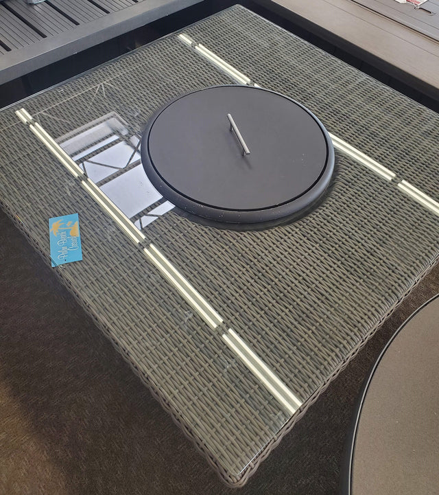Square fire pit table. Made of woven materials. Features a lid. Underneath the lid is fire glass.