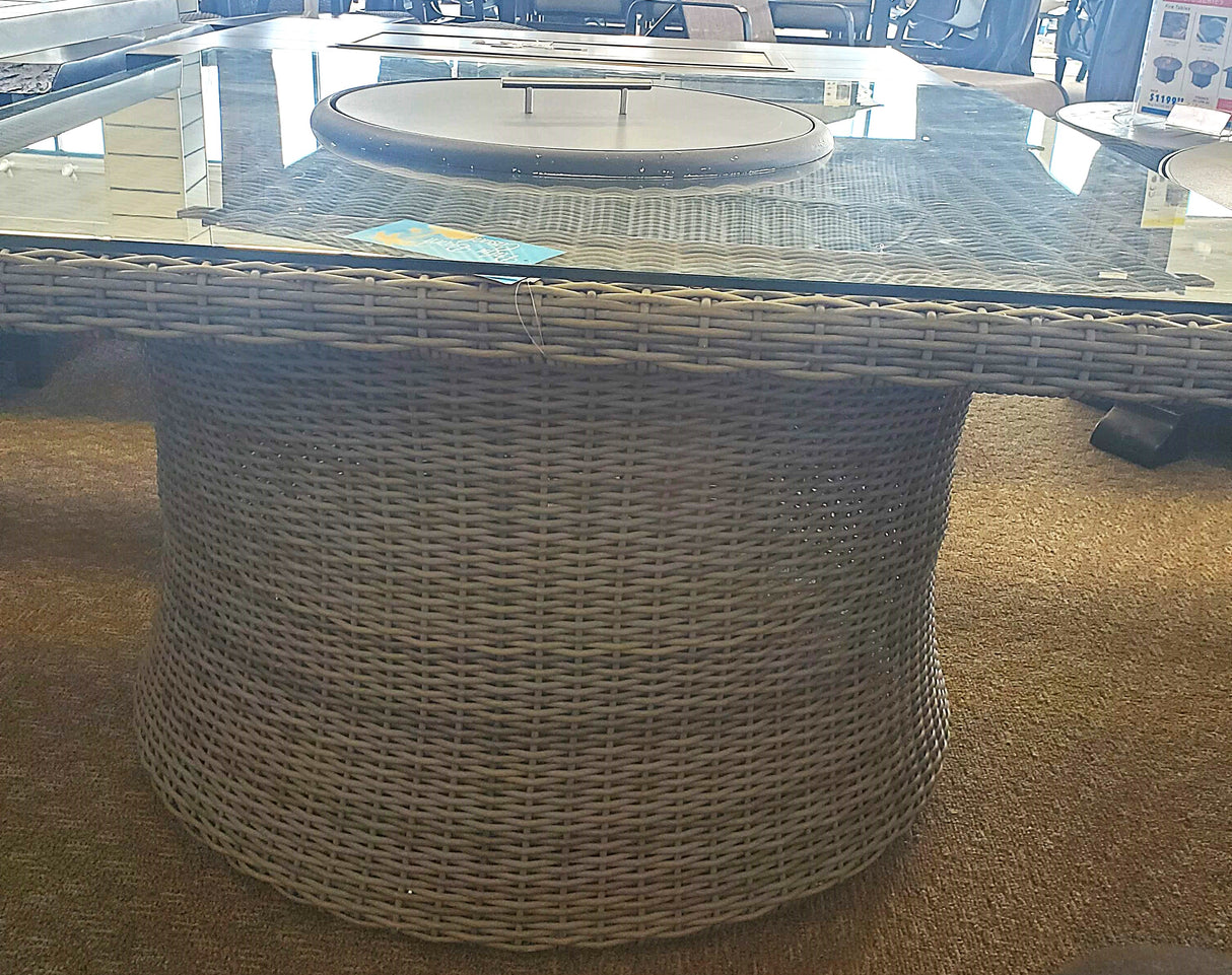 Square fire pit table. Made of woven materials. Features a lid. Underneath the lid is fire glass.