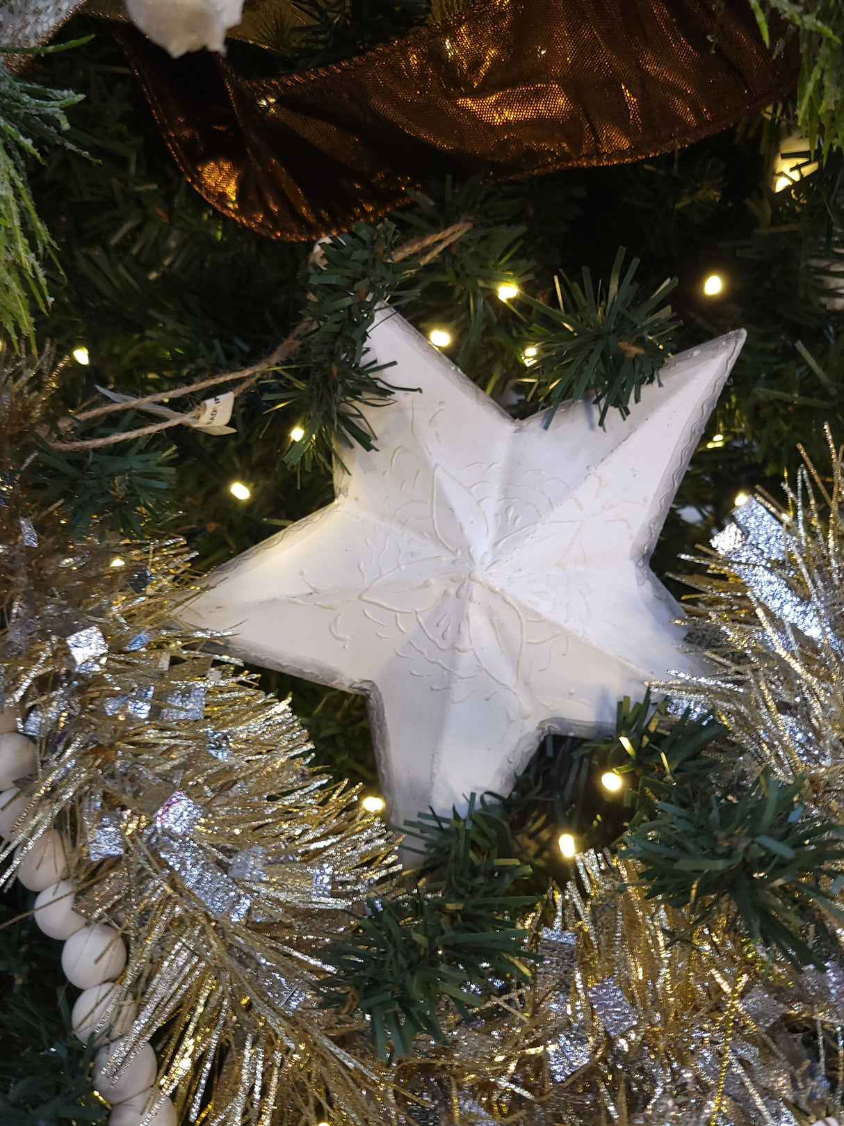 This is a lifestyle image showing the star ornament hanging from a tree branch. It is surrounded by other ornaments, ribbons and beads.