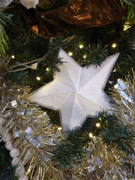 This is a lifestyle image showing the star ornament hanging from a tree branch. It is surrounded by other ornaments, ribbons and beads.