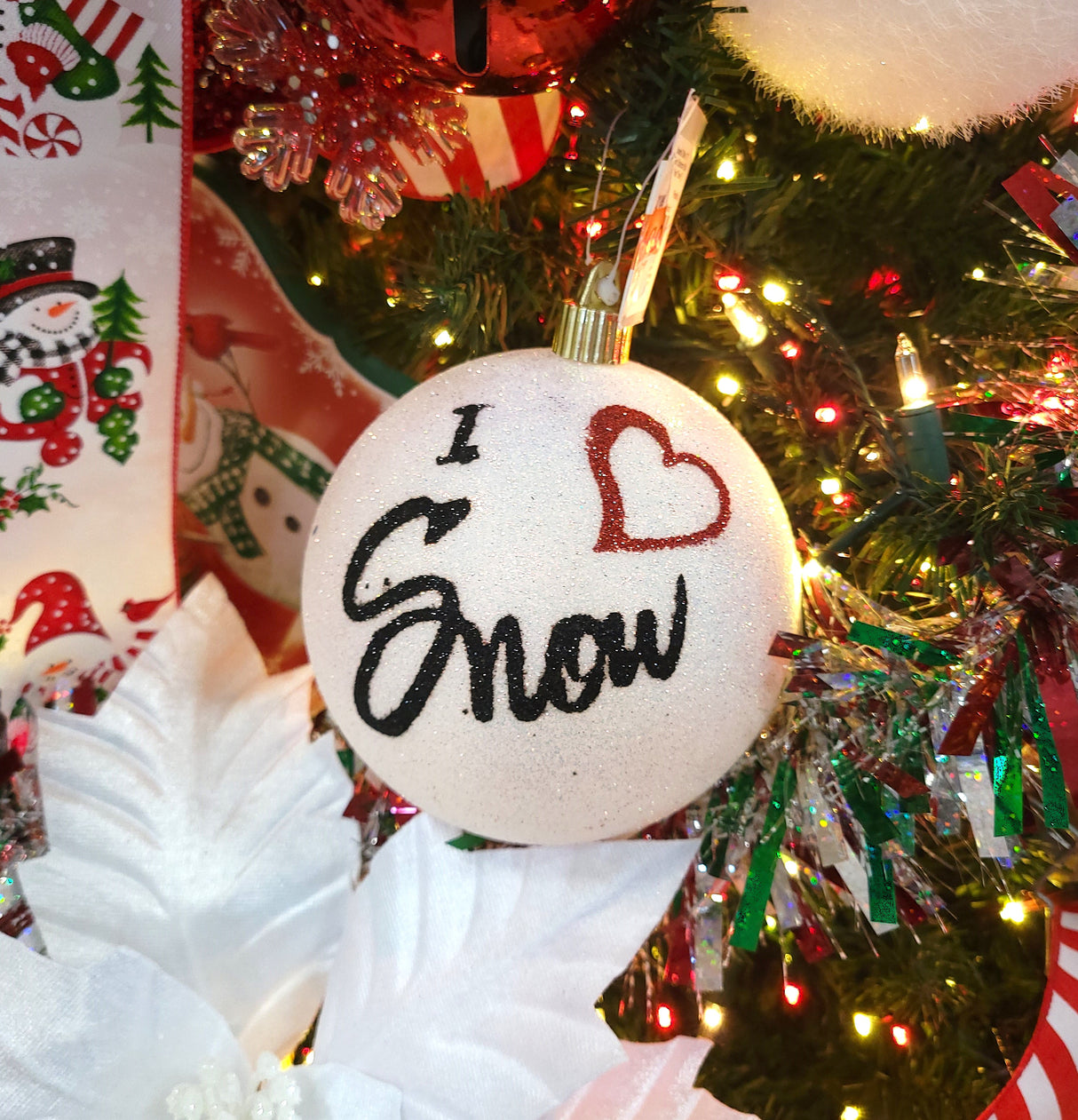 White glittered ornament that reads "I love Snow" in black cursive font. The word "love" is represented by a red glitter heart.