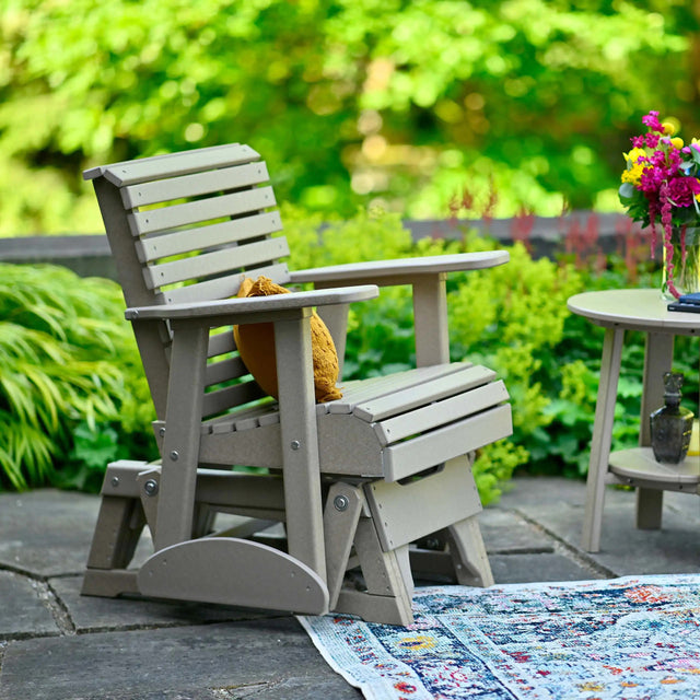 This is a lifestyle image showing off the Weatherwood glider chair on a stone patio. There's a yellow throw pillow on the seat.
