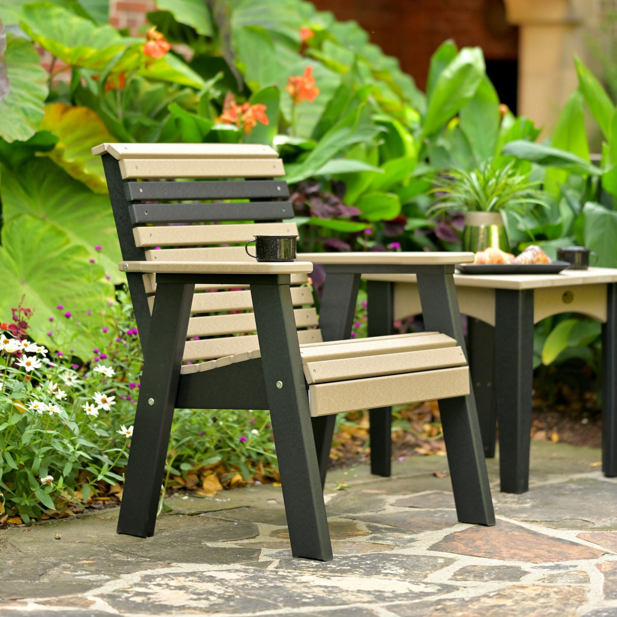 This is a lifestyle image showing the Weatherwood and Black colored bench on a stone patio next to a matching chair. There's a black mug on the armrest.