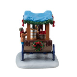 Resin table accent for a Lemax Village tabletop display. Designed to look like a wooden vendor cart. The sign reads "Coffee Wassail." The roof above the carriage is a light blue color and features a green garland around the edges. The vendor is wearing a marron jacket and top hat and is handing a cup to a young child wearing winter clothes. Inside the cart, there's a coffee maker and bowl of wassail warming over a heater. There's a decorated wreath on the back of the cart.