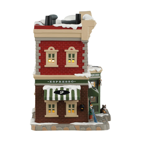 Porcelain lighted building for a Lemax tabletop display. Designed to look like a red, gray and brown brick building. There's snow on the roof and the sidewalk. There are signs advertising croissants, coffee, espresso and sandwiches. Inside the window you can see a man sitting at a table. There's a dog sitting under the window on the sidewalk.